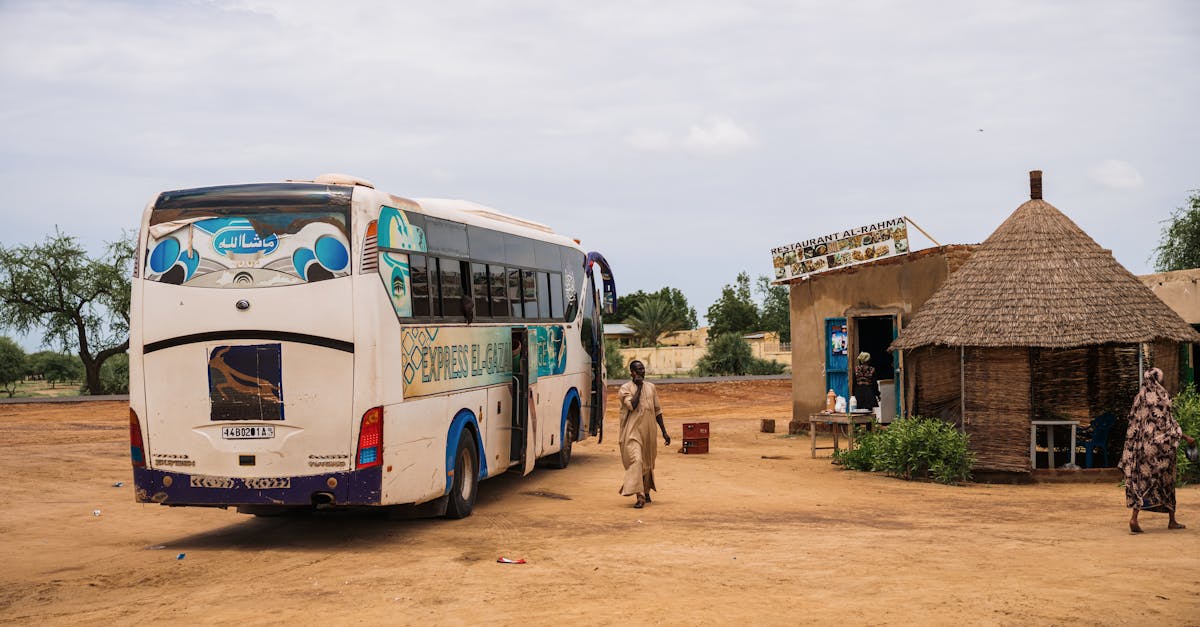 A white bus stops at a rural village with a tribal hut and local restaurant.